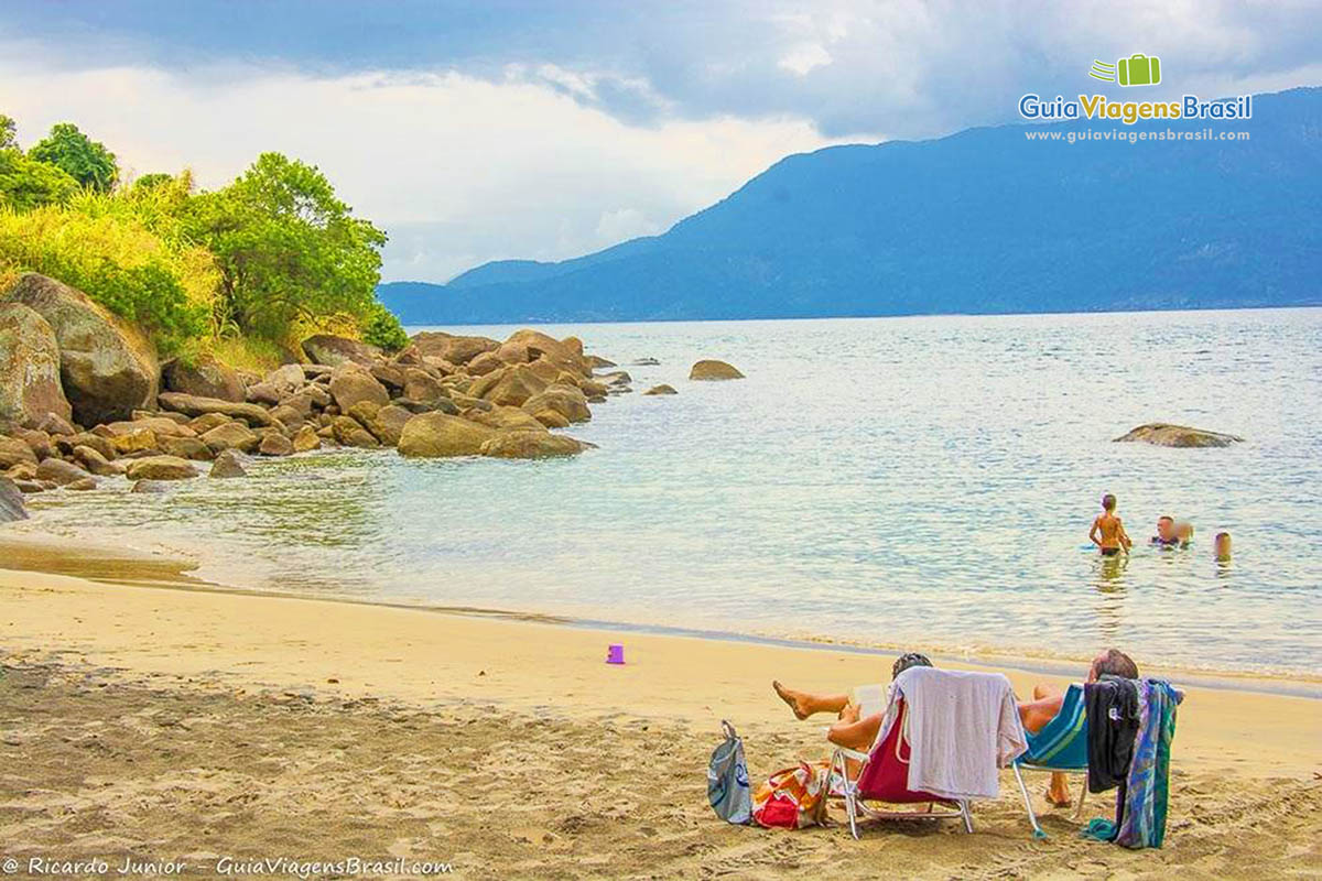 Imagem de casal sentados na areia e pessoas nas águas da praia.