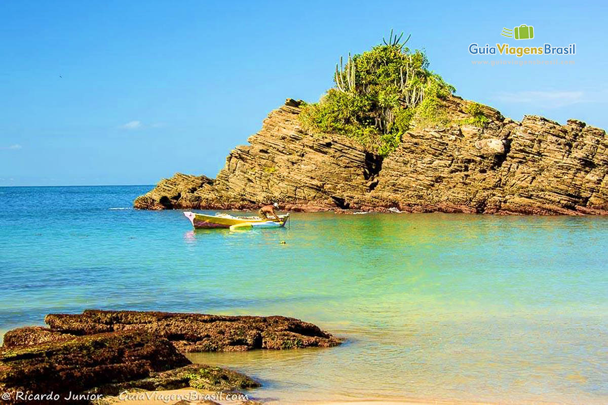 Imagem de mar cristalino e ao fundo pedras da praia. 