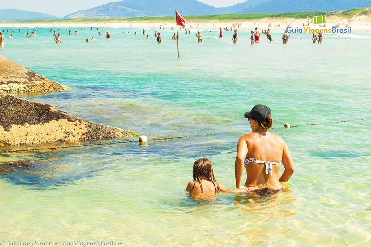 Imagem de mãe e filha se molhando nas águas da Praia Joaquina. 