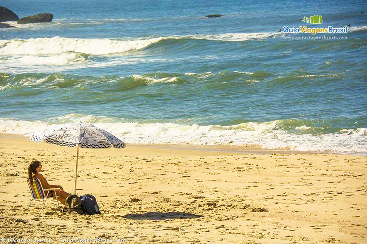 Imagem de uma menina sentada em baixo de um guarda sol na Prainha, curtindo um belo dia de praia.