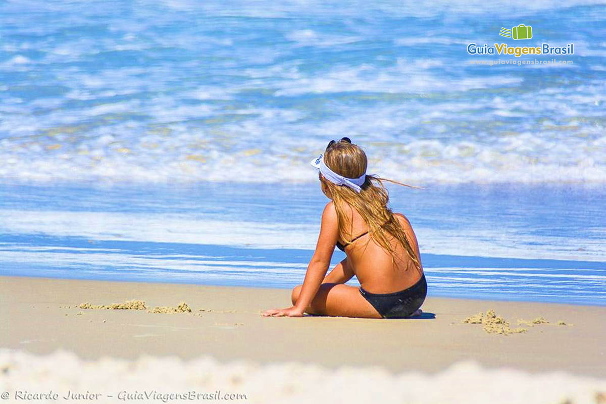 Imagem de uma menina sentada na areia admirando o mar.
