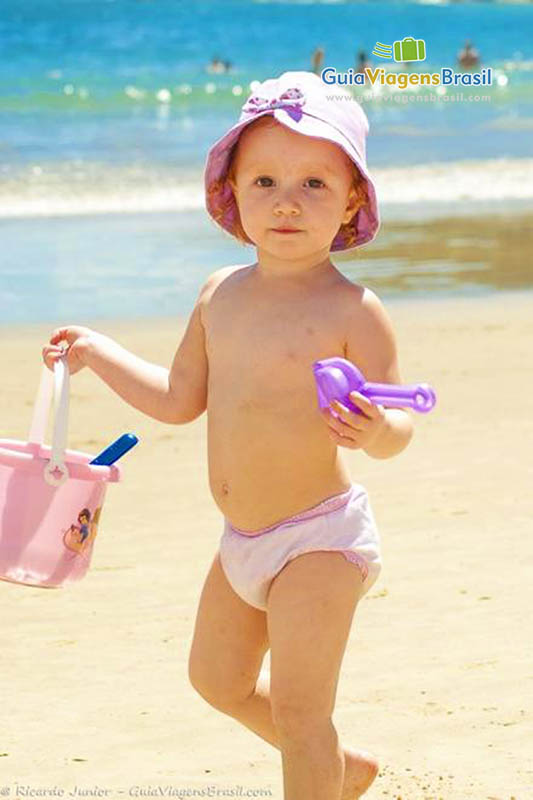 Imagem de uma menininha na areia com seu baldinho de brincar.