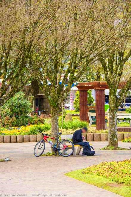 Imagem de menino sentado no banco da praça com sua bicicleta ao lado.