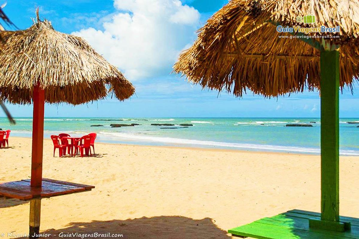 Imagem das mesas de madeira e guarda sol de sapê nas areias da praia.