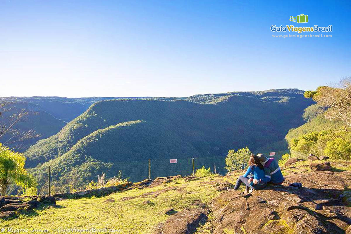 Imagem de um casal sentados em uma grande pedra vendo no horizonte os belos morros.