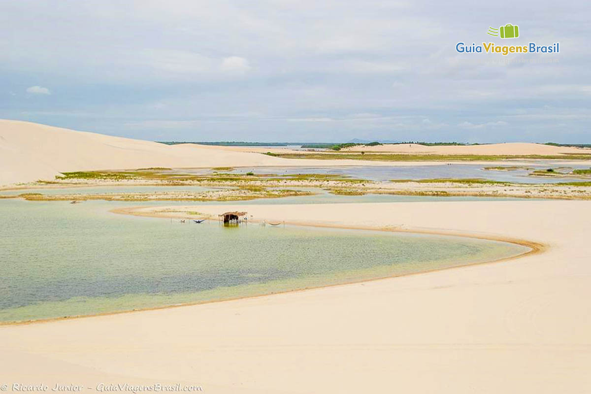 Imagem das dunas, de um lago e uma barraca de sapê na beira do pequeno lago.