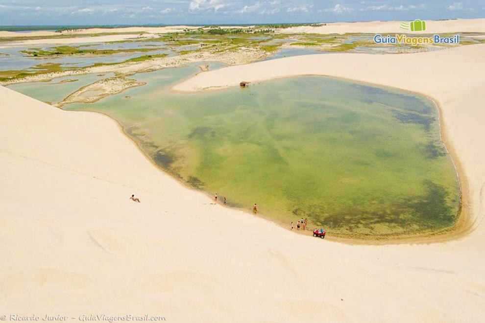 Imagem de um grande lago com turistas na beira do lago e outros escorregando nas dunas até o lago.