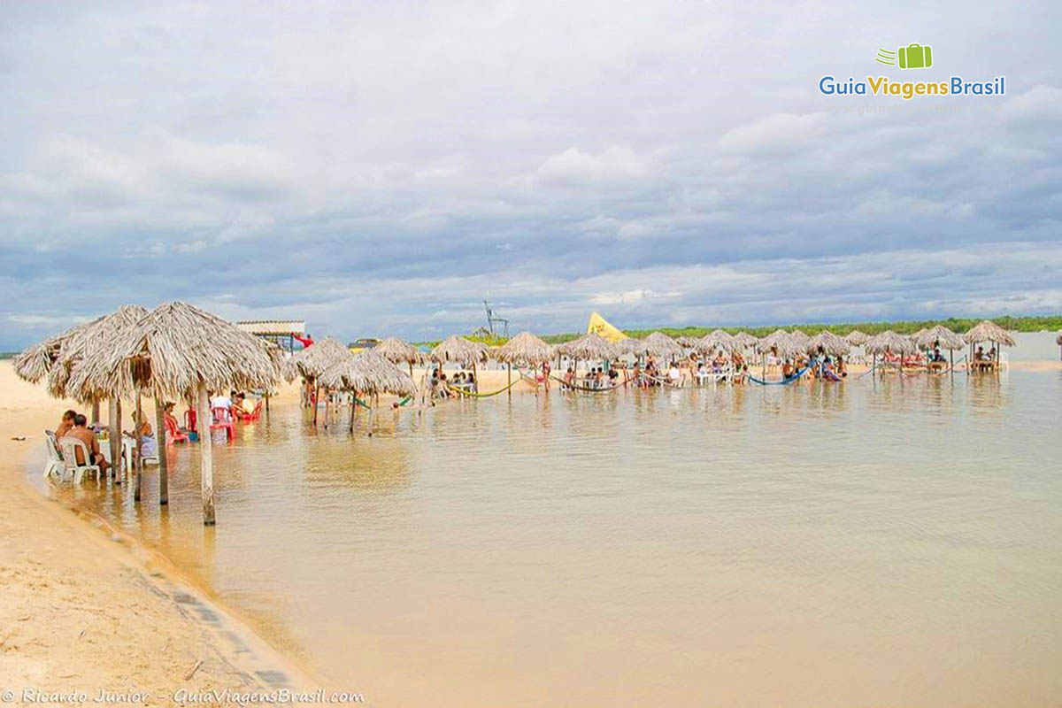 Imagem de vários guarda sol de sapê em volta do lago, com turistas aproveitando dia de sol.