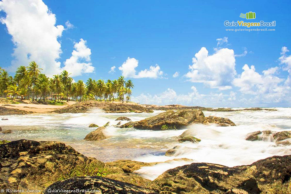 Imagem de águas batendo nas pedras da lindíssima praia.