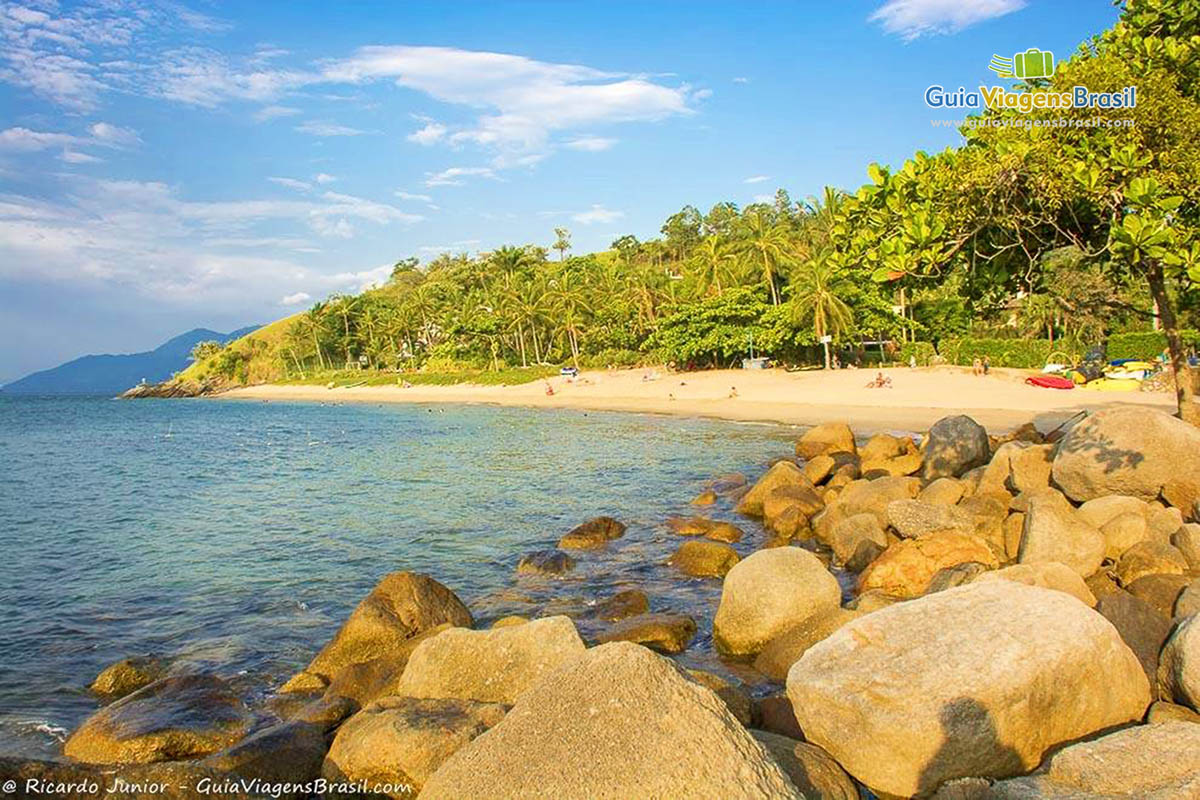 Imagem das pedras, do mar e da orla repleta de vegetação na Praia da Feiticeira. 