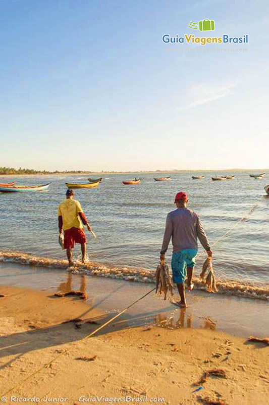 Imagem de pescadores na beira do mar com peixes na mão e ao fundo no mar vários barcos de pescador.