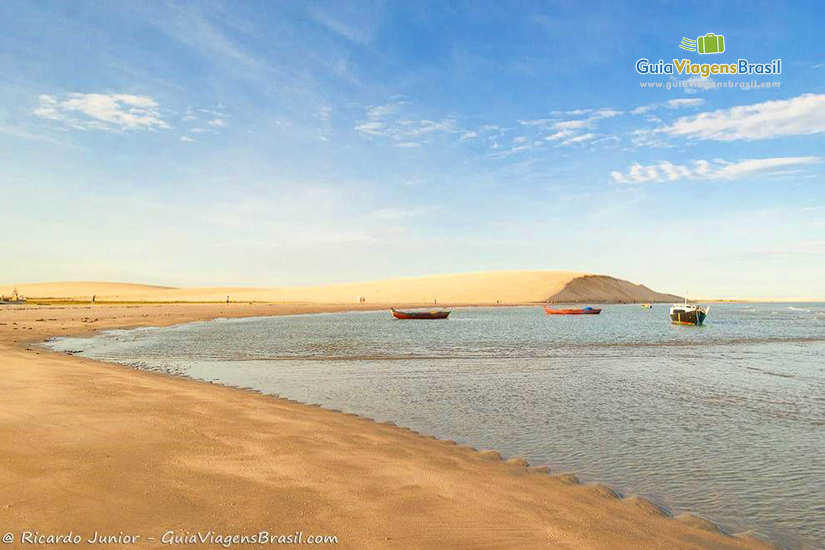 Imagem de pequenos barcos de pescador parado na água próximo as dunas.