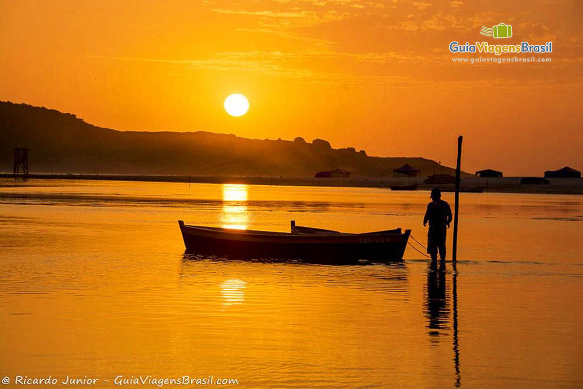 Imagem de uma pessoas ao lado de um barco de pescador na beira da praia, admirando o  magnífico entardecer.