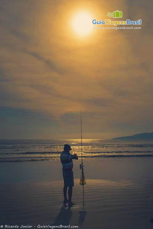 Imagem de um pescador com vara ao lado na beira da praia.