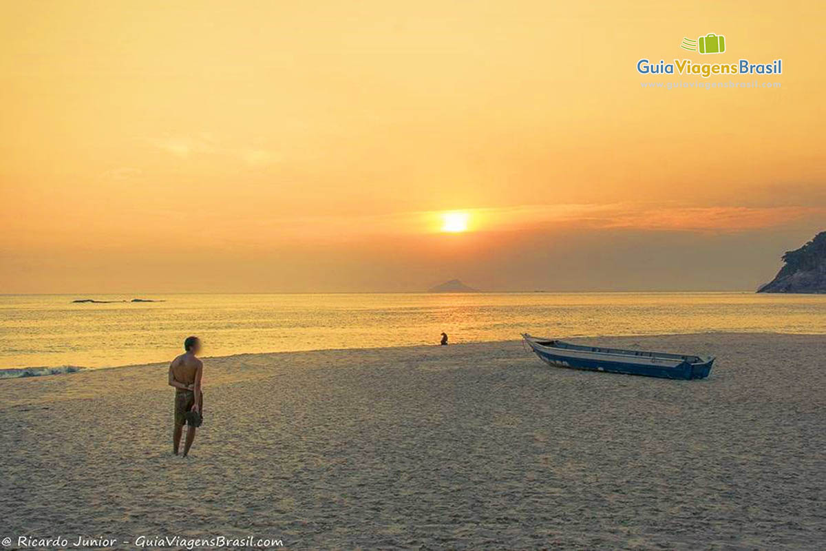 Imagem de um rapaz admirando o por do sol e um cachorro sentado próximo ao mar olhando para sol no horizonte.