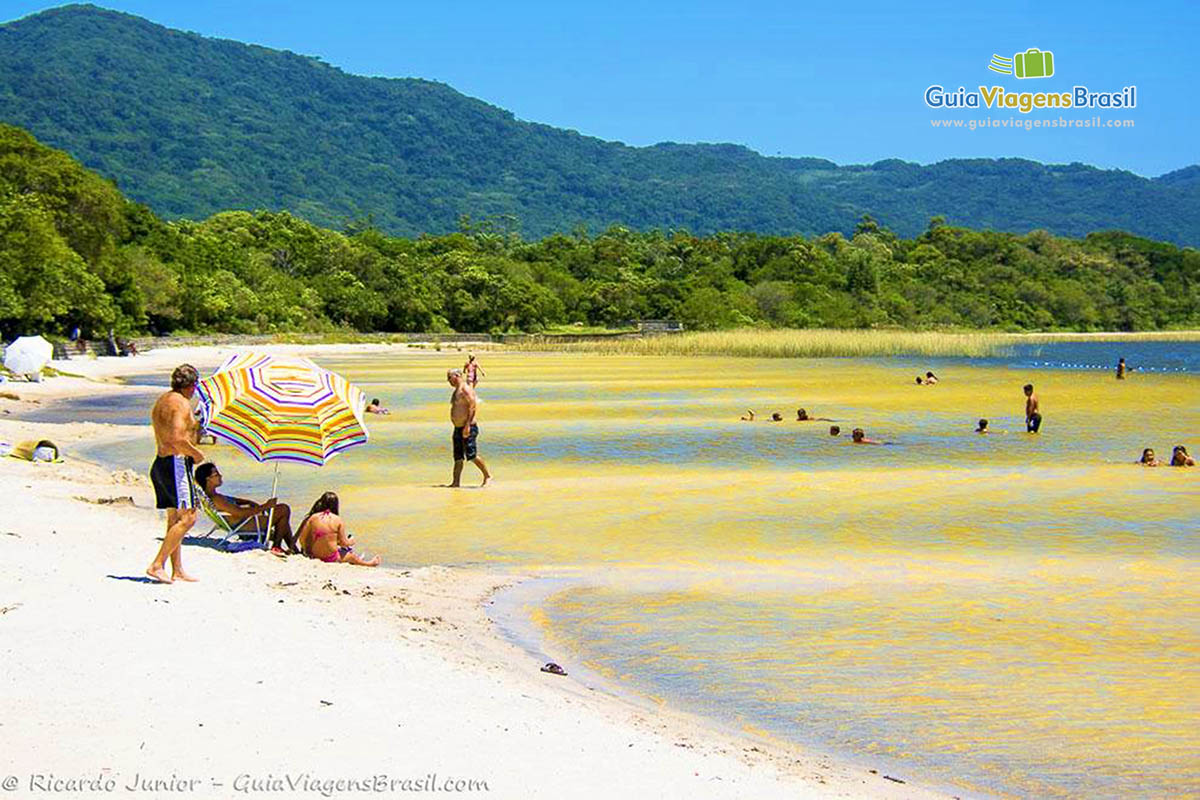 Imagem de família em baixo de guarda sol próximo a lagoa.