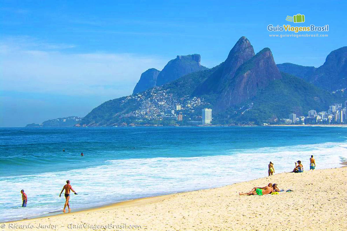 Imagem de pessoas caminhando na beira da praia e pessoas tomando sol nas areias.