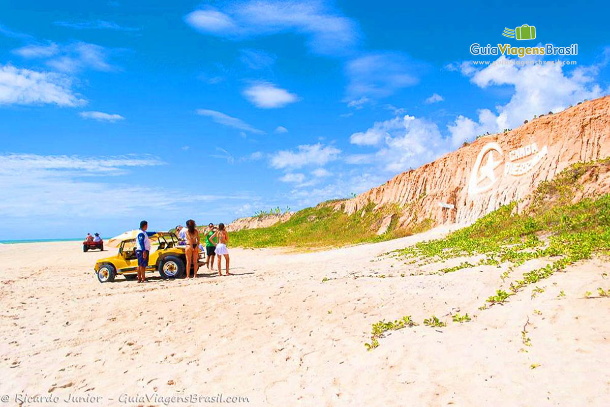 Imagem de bugue parado com turistas tirando foto do símbolo da praia esculpido na falesia. 