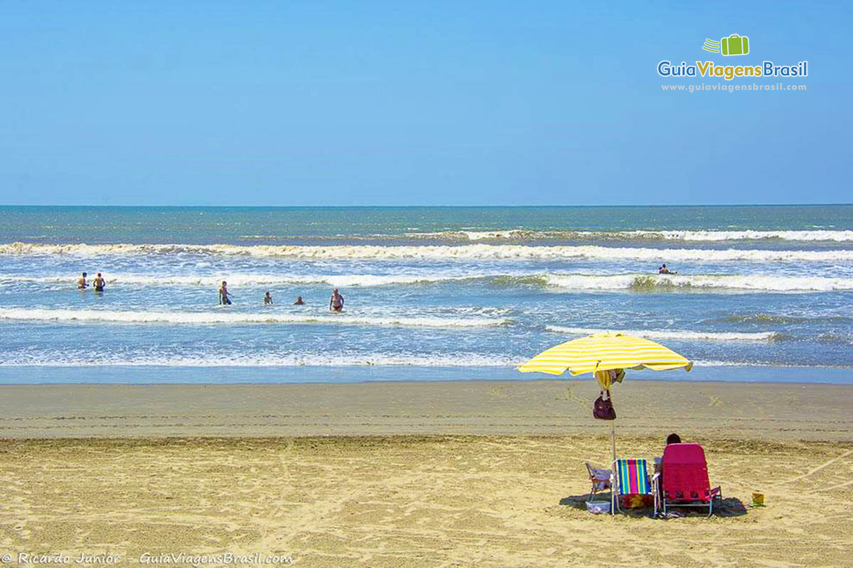 Imagem de um guarda sol amarelo na areia e nas águas as pessoa aproveitando.