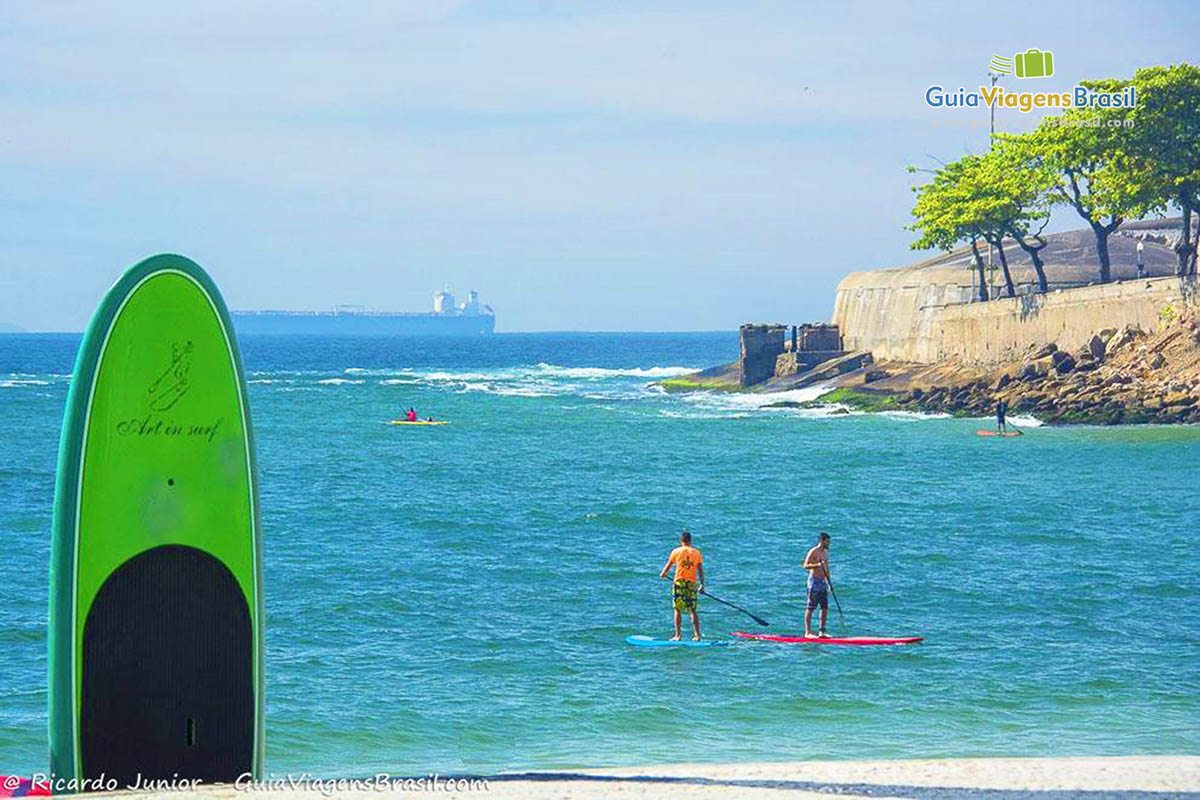 Imagem da prancha de stand up paddle e ao fundo pessoas nas águas praticando o badalado esporte.