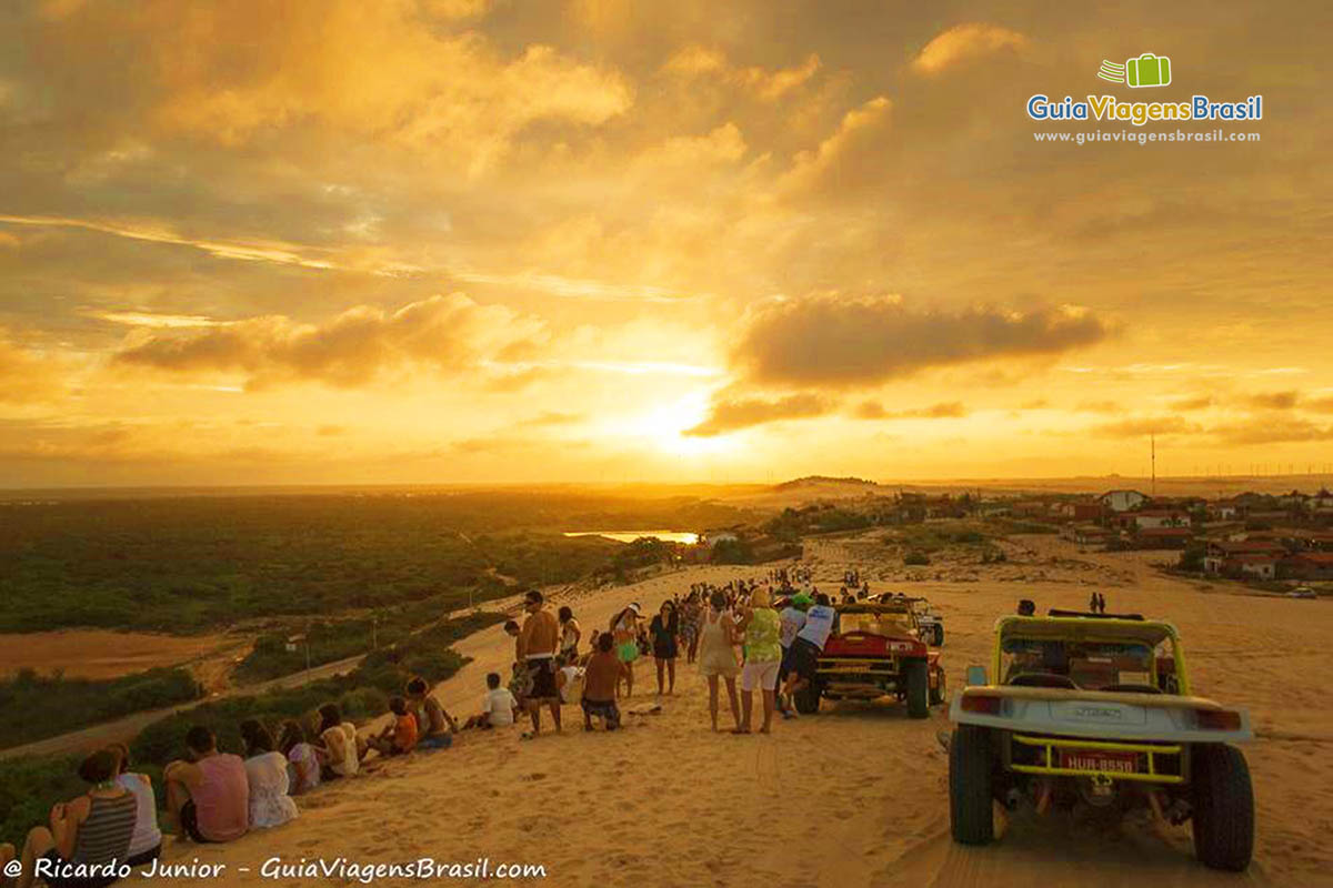 Imagem de turistas sentados nas Dunas para admirar um belo pôr do sol.