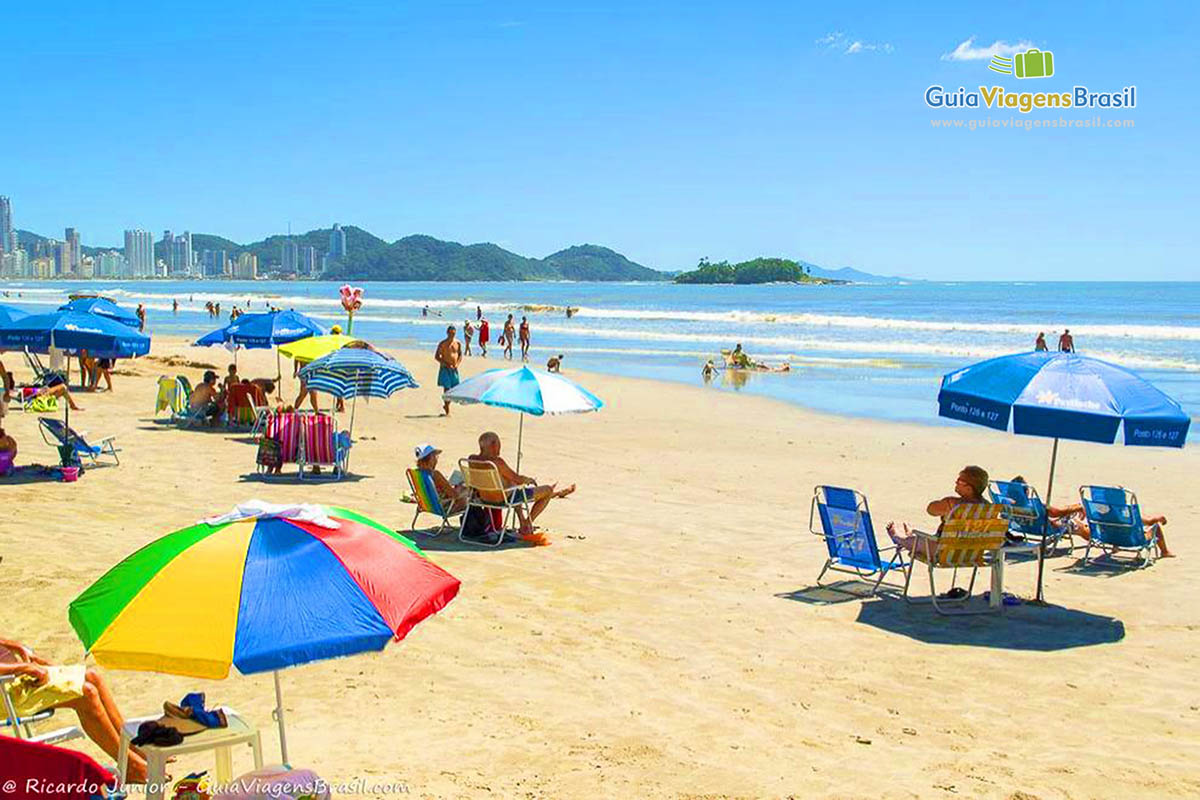 Imagem de turistas com guarda sol coloridos nas areias da praia.