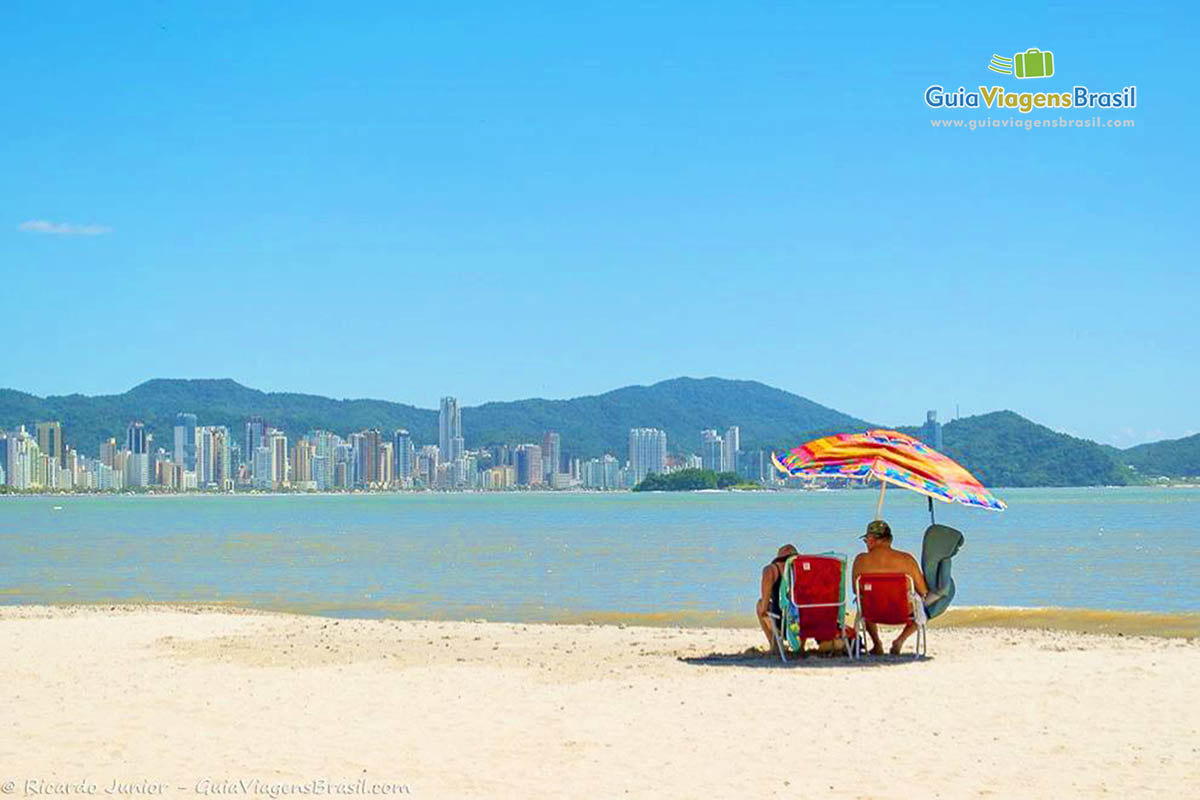 Imagem de um casal sentados embaixo de guarda sol, olhando o lindo mar.