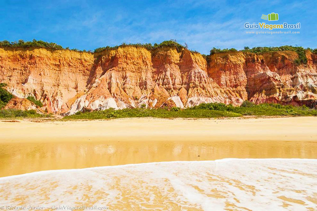 Imagem das águas na beira da praia e ao fundo falesia avermelhada.