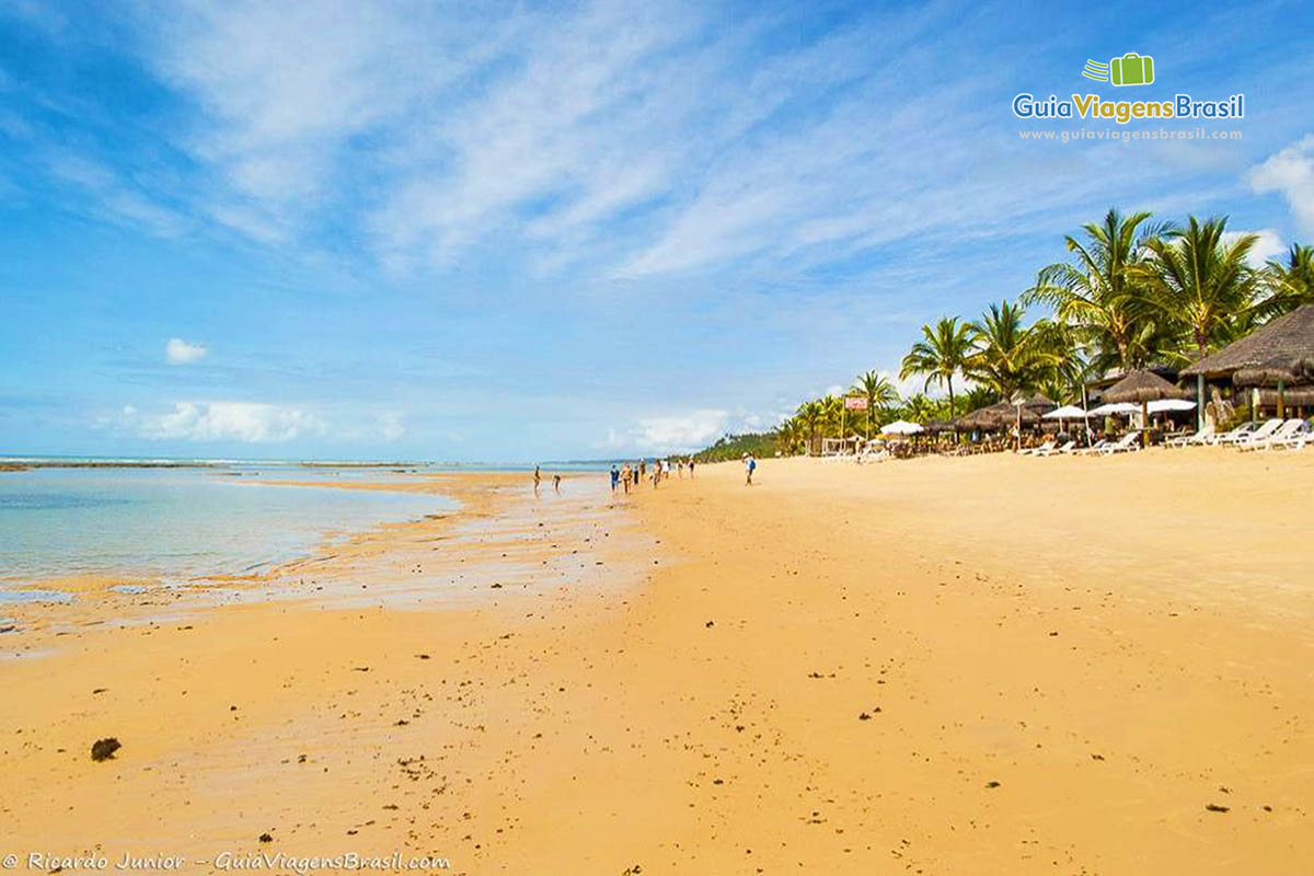 Imagem de quiosques na orla e de turistas tirando fotos na areia.