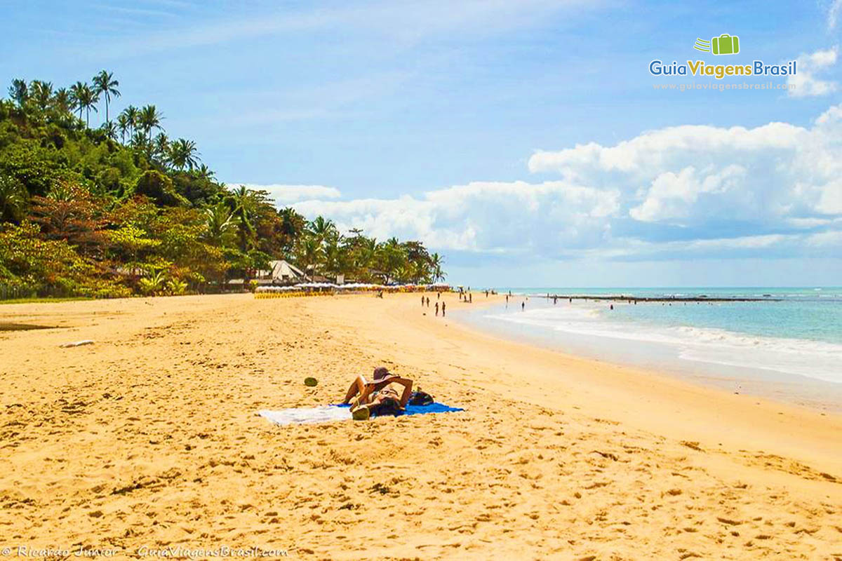 Imagem de turistas tomando sol ao fundo o mar e recifes na Praia Pitinga.