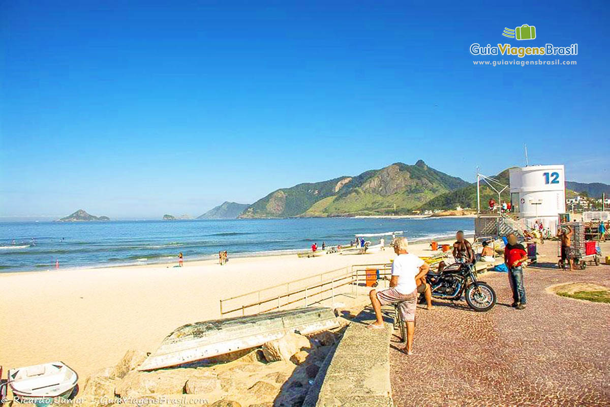 Imagem de homens no calçadão ao lado do Posto 12 admirando o mar da praia.