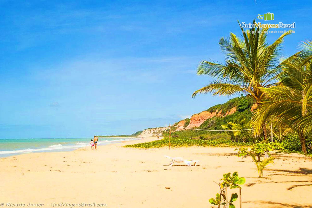 Imagem de uma rede de vôlei na areia e pai e filha andando nas areias da Praia Taipe.