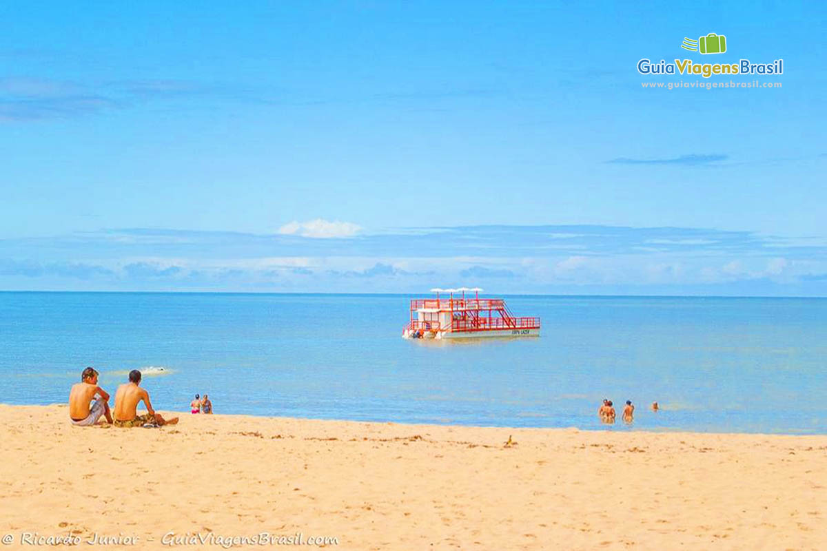 Imagem de dois rapazes sentados na areia, conversando e admirando a bela vista da praia.