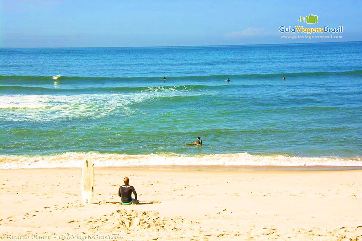 Imagem de um sufista sentado nas areias e outros no mar aguardando ondas.