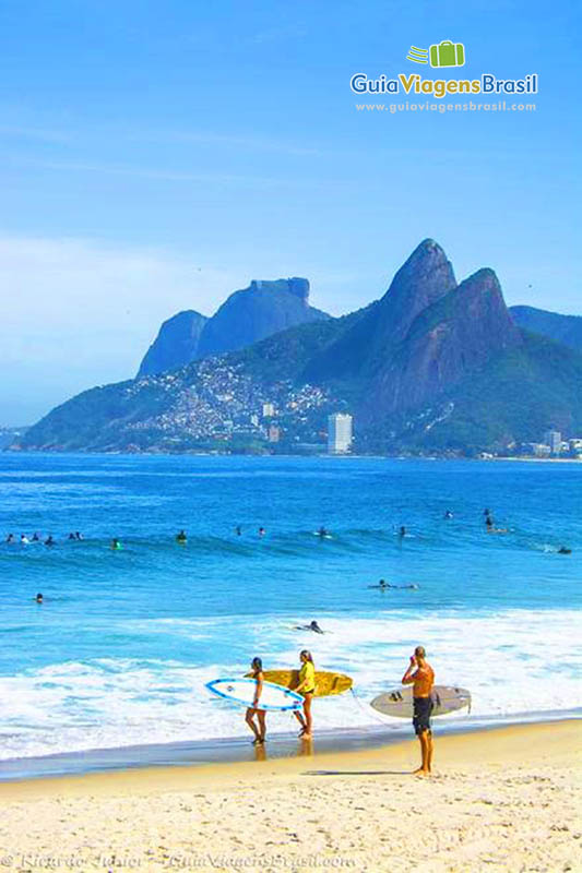 Imagem de três surfistas na beira da praia segurando suas pranchas e olhando para mar.