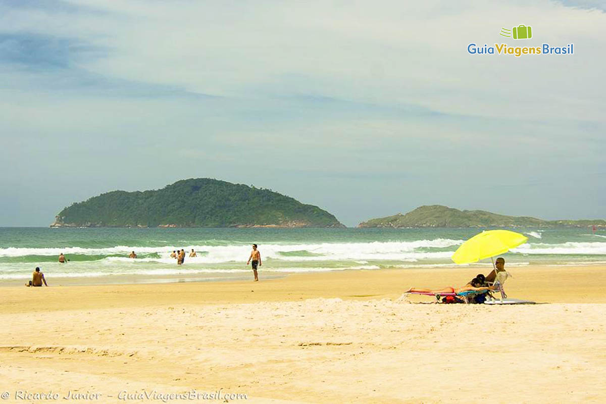 Imagem de um guarda sol amarelo na areia com pessoa embaixo se protegendo do sol.